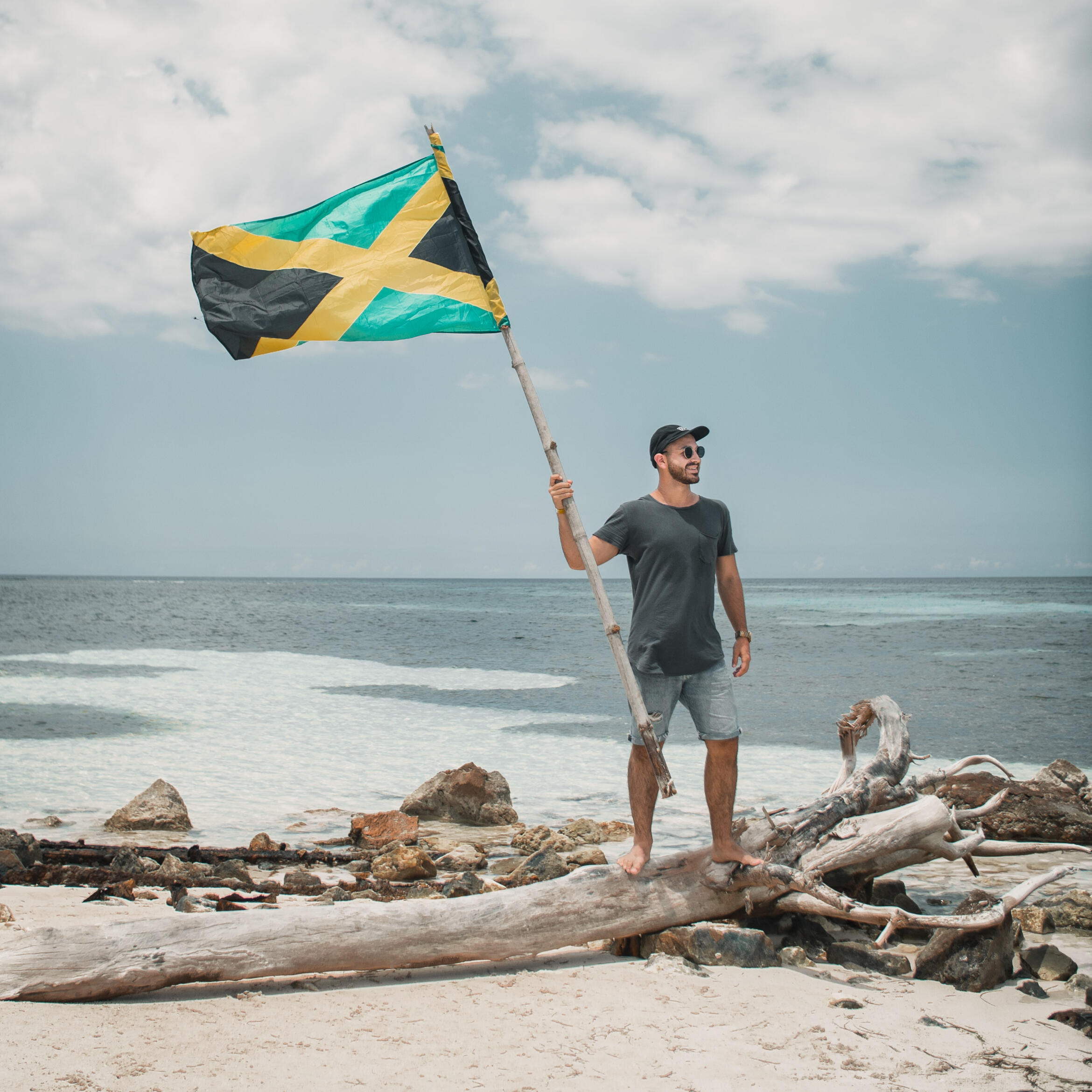 A white man holding a Jamaican flag while standing on top of a huge piece of driftwood